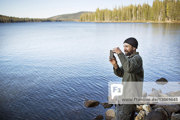 Mann fotografiert mit Handy am Seeufer stehend