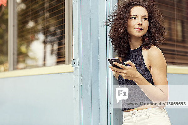 Frau mit Smartphone schaut weg  während sie am Gebäude steht