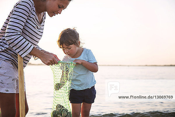 Mutter zeigt Tochter Muscheln im Fischernetz am Strand vor klarem Himmel