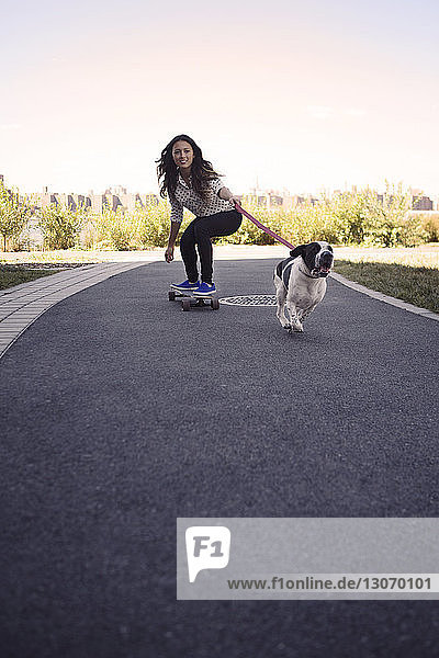 Frau fährt Skateboard mit laufendem Hund auf der Straße gegen den klaren Himmel