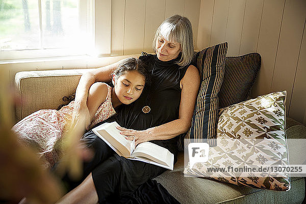 Grandmother reading book to granddaughter while resting on couch at home