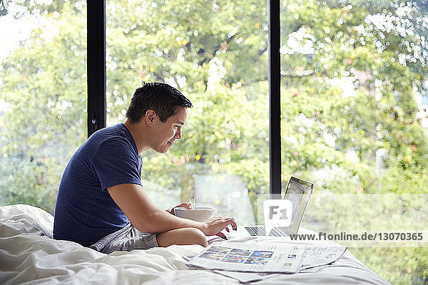 Seitenansicht eines Mannes mit Laptop beim Frühstück am Bett am Fenster