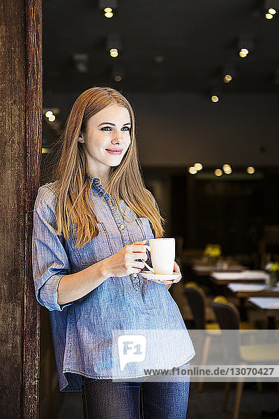 Thoughtful smiling woman holding coffee cup while standing at cafe
