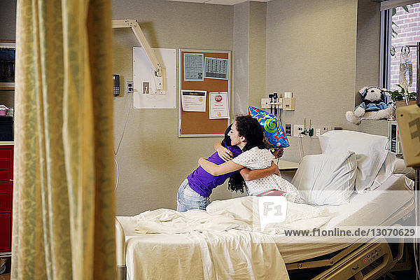 Mother embracing girl while sitting on bed in hospital