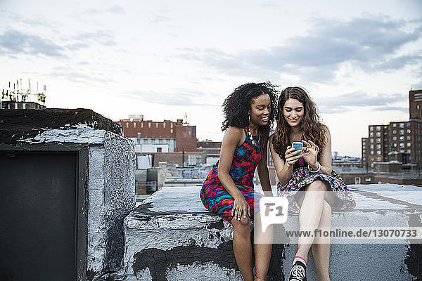 Freunde benutzen Mobiltelefon  während sie auf der Gebäudeterrasse gegen den Himmel sitzen