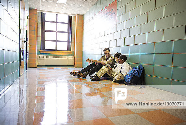 Boys using tablet computer while sitting with teacher in corridor