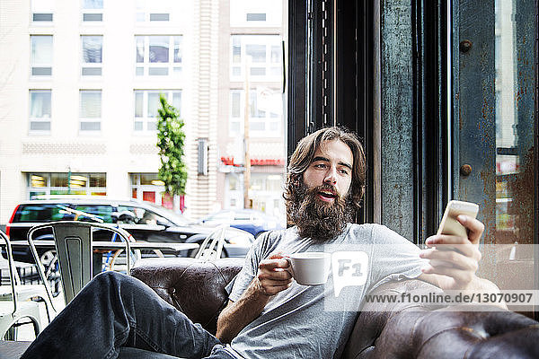 Man having coffee and using phone while sitting against window at cafe
