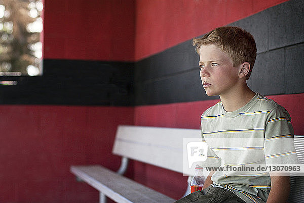 Boy looking away while sitting on bench at dugout