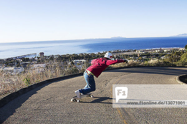 Rear view of man longboard skating on road in town by sea against clear sky