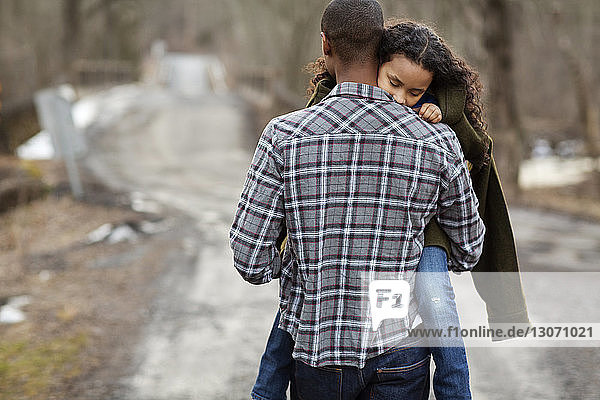 Rückansicht eines Vaters  der seine Tochter trägt  während er auf einer Straße im Wald geht