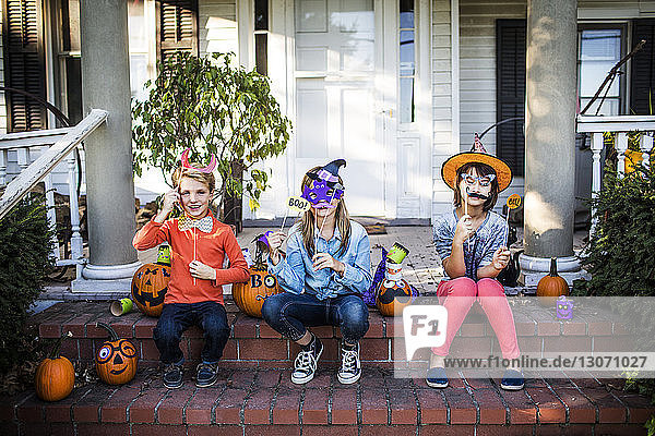 Glückliche Geschwister halten Requisiten und sitzen an Halloween mit geschmückten Kürbissen auf den Stufen