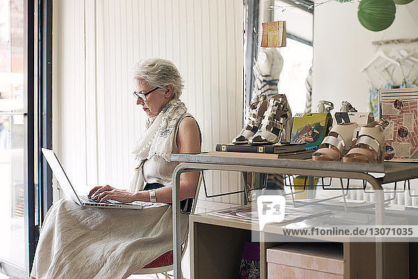 Seitenansicht einer älteren Frau  die einen Laptop-Computer benutzt  während sie auf einem Stuhl im Laden sitzt