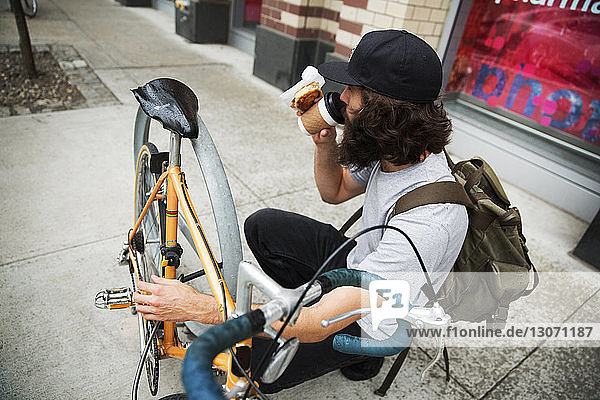 Schrägaufnahme eines Mannes  der Kaffee trinkt  während er mit dem Fahrrad auf dem Bürgersteig kauert