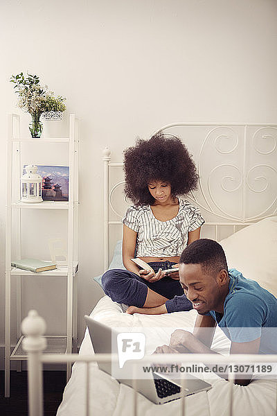 Smiling man using laptop computer while woman reading book on bed at home