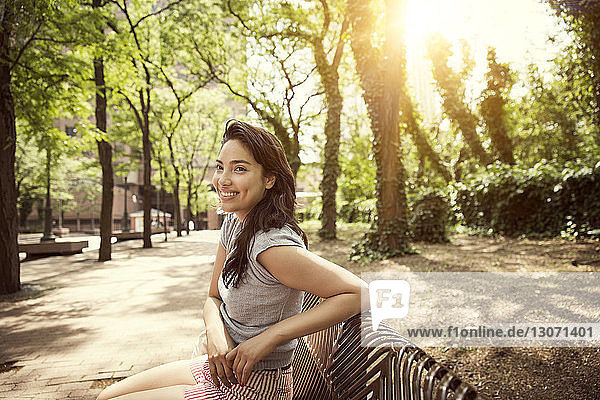 Lächelnde Frau sitzt an sonnigen Tagen im Park auf Bank an Bäumen