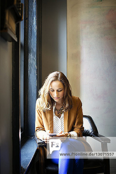 Frau benutzt Smartphone  während sie im Büro am Fenster sitzt