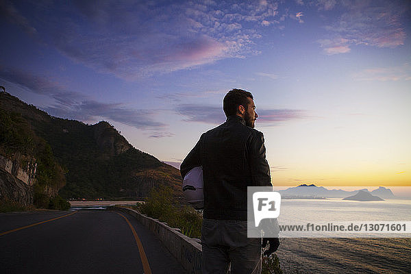 Rückansicht eines Mannes mit Helm  der bei Sonnenuntergang auf der Küstenstraße steht