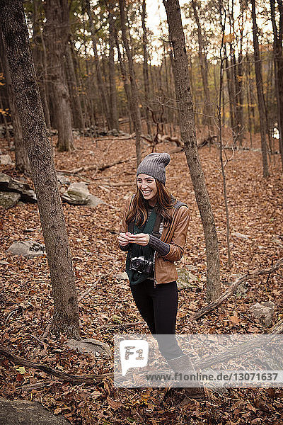 Smiling woman with camera standing amidst trees in forest during winter