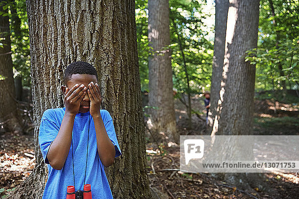 Verspielter Junge  der die Augen bedeckt  während er im Wald an einem Baum steht