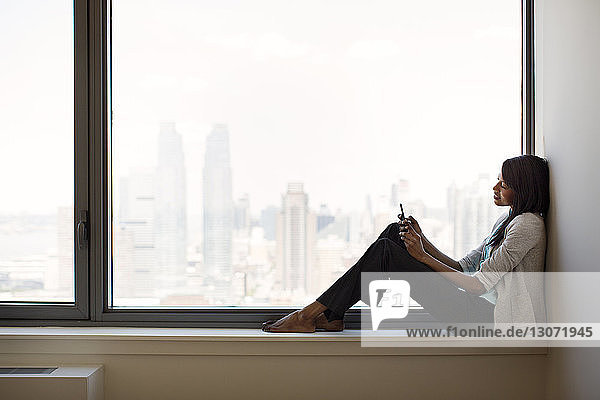 Geschäftsfrau benutzt Mobiltelefon  während sie im Büro auf dem Fensterbrett sitzt