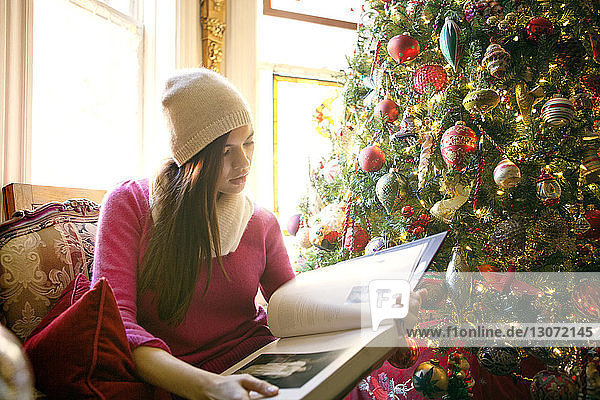 Frau liest Buch  während sie zu Hause am Weihnachtsbaum sitzt