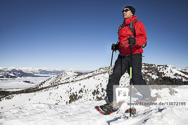 Man with ski poles walking on snow covered field against clear blue sky