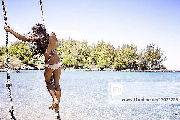 Rückansicht einer Frau im Bikini  die am Strand am Seil schwingt