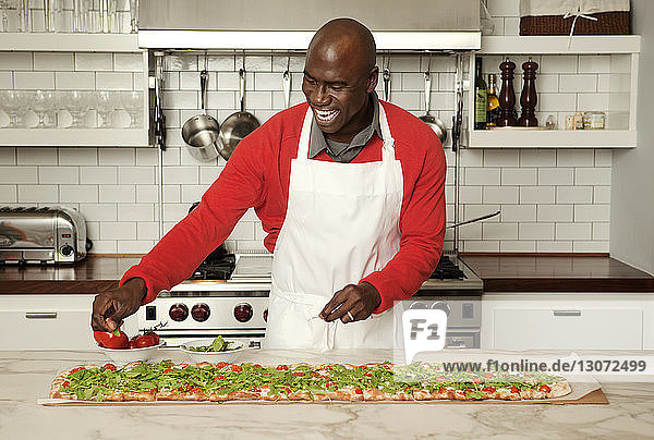 Fröhlicher Mann garniert Pizza auf Kücheninsel zu Hause