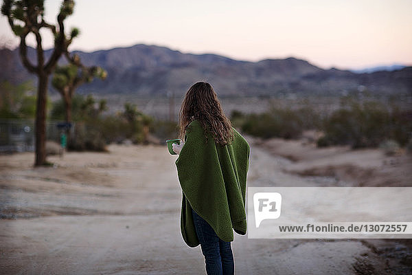 Frau in eine Decke gewickelt  die eine Kaffeetasse hält  während sie auf einem Fußweg steht