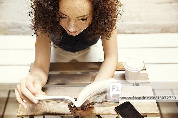 Hochwinkelansicht einer Frau  die ein Buch liest  während sie in einem Cafe auf dem Bürgersteig sitzt