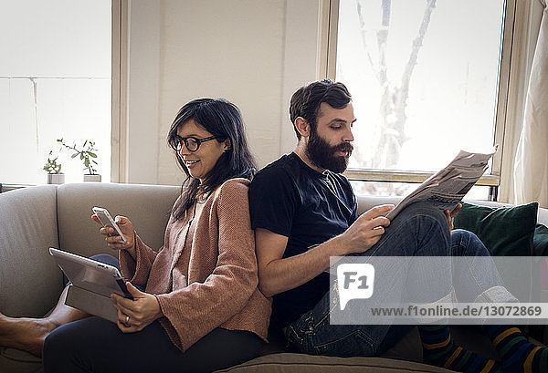 Woman using mobile phone while husband reading newspaper on sofa