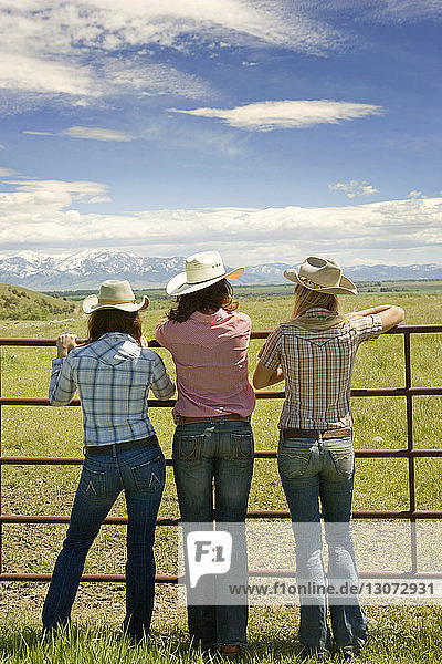Rear view of cowgirls standing by gate against sky