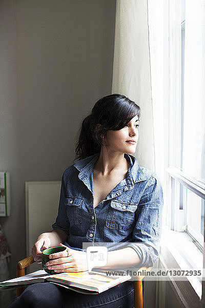 Woman looking away while sitting on chair by window at home