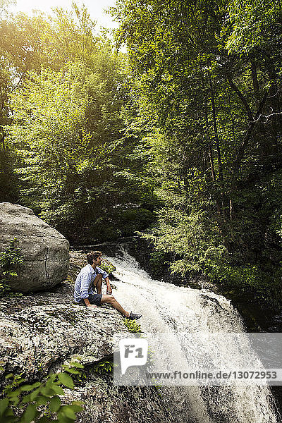 Side view of man sitting on rocks by waterfall in forest