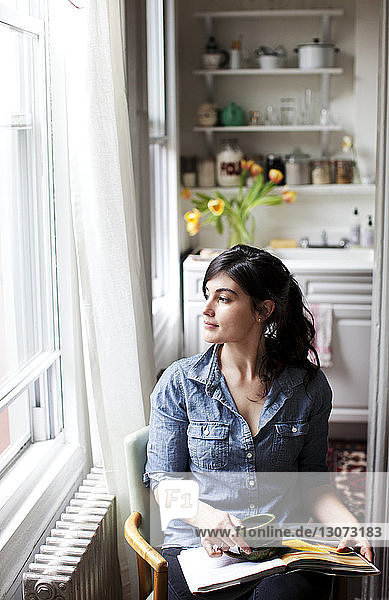 Woman looking away while sitting on chair by window at home