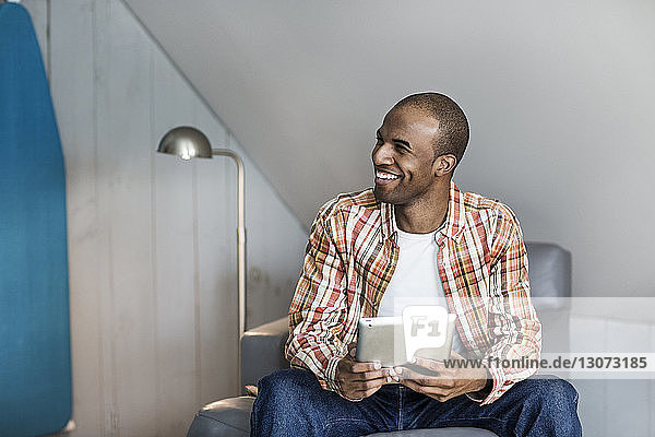 Man with tablet computer looking away while sitting on chair at home