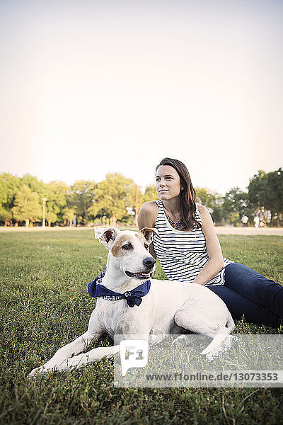 Thoughtful woman sitting with dog on grassy field against clear sky at park