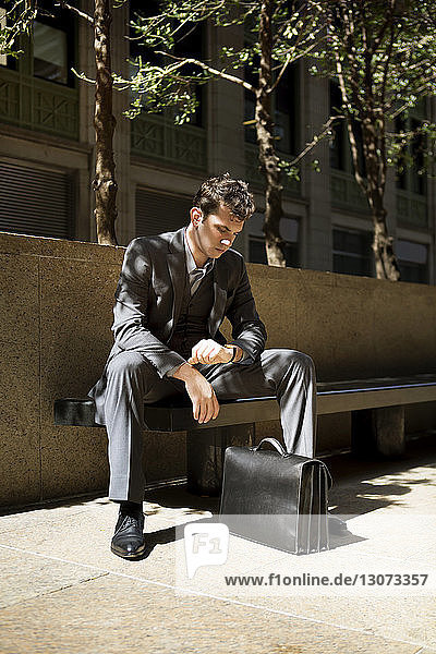 Mann mit Aktentasche schaut auf die Armbanduhr  während er auf einer Bank gegen das Gebäude sitzt
