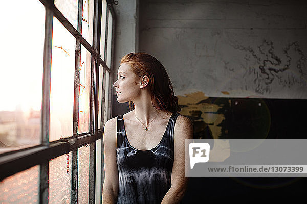 Thoughtful woman looking through glass window while standing against wall