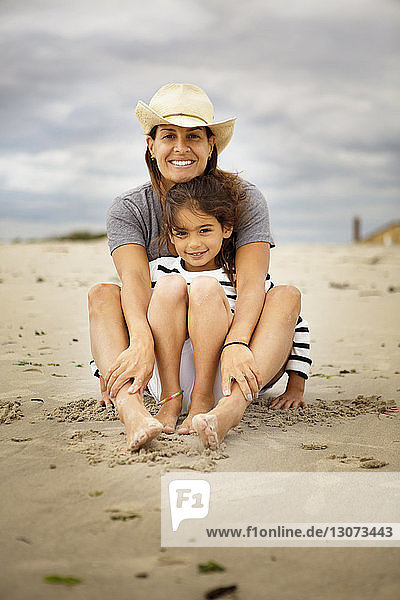 Portrait of smiling mother and daughter sitting on sand against cloudy sky
