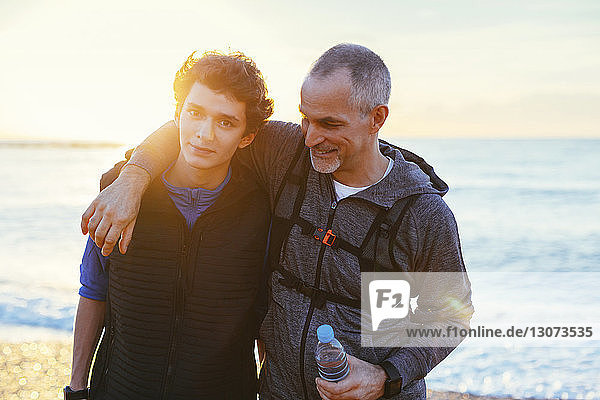 Portrait of son with father's arm around shoulder against sea during sunset