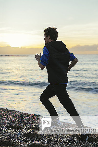 Full length of teenage boy jogging on shore at beach against sky during sunset