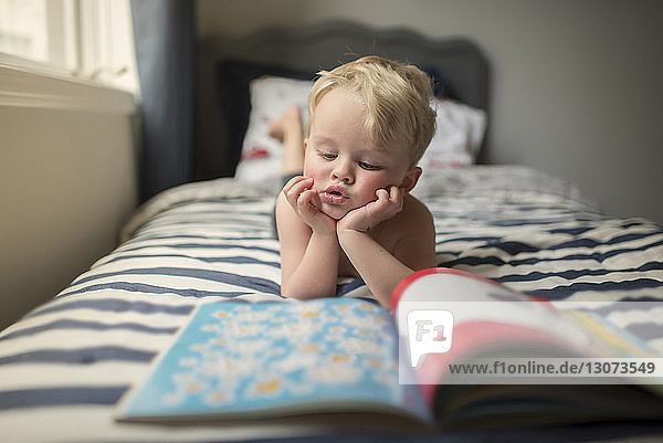 Junge mit Händen am Kinn liest Bilderbuch  während er zu Hause auf dem Bett liegt