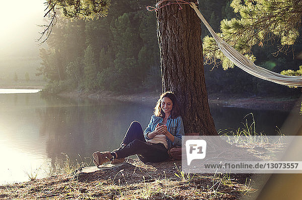 Happy woman using mobile phone while relaxing at lakeshore