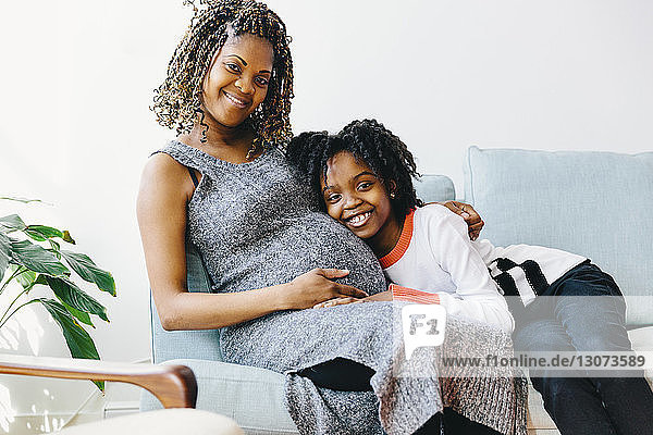 Porträt einer glücklichen Tochter  die die schwangere Mutter umarmt  während sie zu Hause auf dem Sofa sitzt