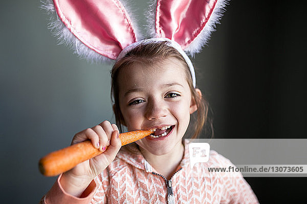 Porträt eines verspielten Mädchens mit Kaninchenohren-Stirnband  das zu Hause in Karotten beißt