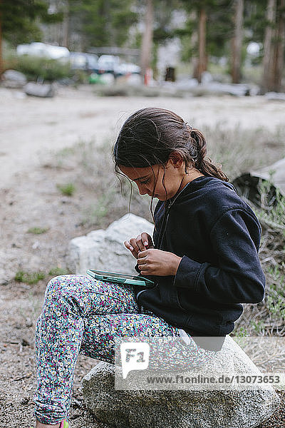Mädchen schaut auf Smartphone  während sie auf einem Felsen auf dem Campingplatz sitzt