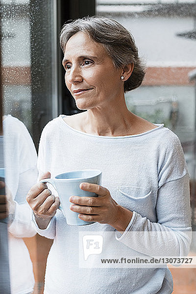 Nachdenkliche reife Frau hält Kaffeetasse  während sie zu Hause durchs Fenster schaut