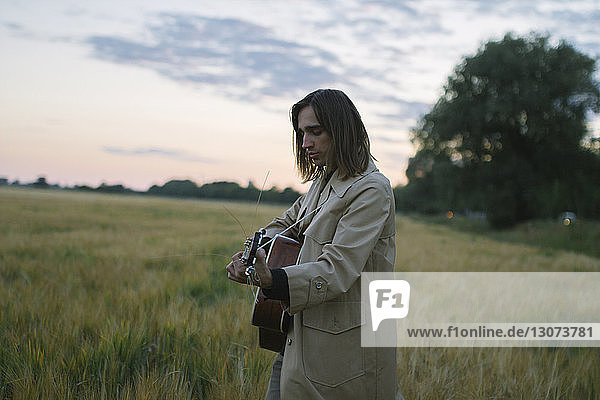 Side view of man playing guitar while standing on grassy field against sky