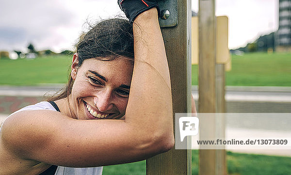 Glückliche Frau schaut weg  während sie sich im Park an eine Gymnastikstange lehnt
